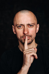 Portrait of a young bald man with stubble, pressing a finger to his mouth, with which he calls for...