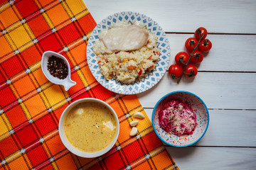 Delicious lunch set contains soup, beetroot and cheese salad, fried chicken breast and rice. Tasty meal is served on the white wooden table covered with orange towel. 