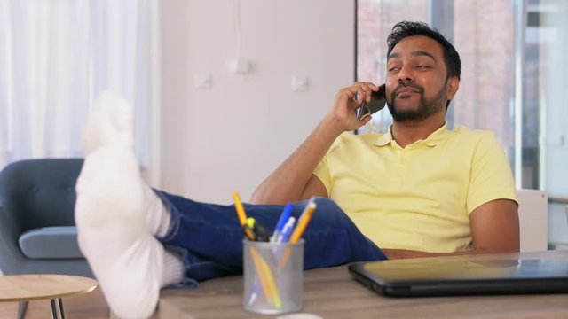 technology, communication and people concept - happy smiling indian man calling on smartphone and resting feet on table at home office