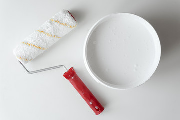 A bucket with white paint and a paint roller on a white table. Repair concept. Top view.