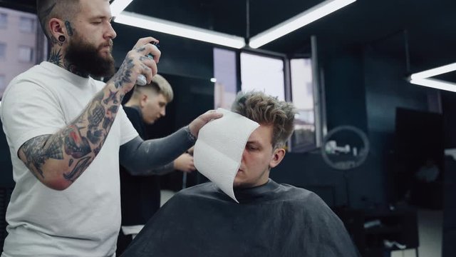 Attractive male is getting a modern haircut in barber shop. Barber dyes hair to the client with a blue spray.