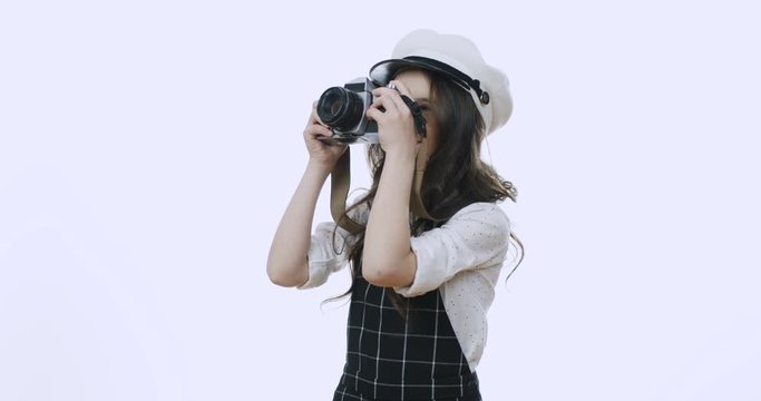 Pretty and cute small stylish Caucasian girl in a hat taking photos with a photocamera and smiling joyfully on the white wall background.