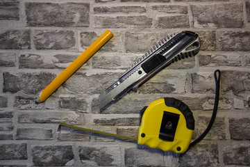 Construction yellow tape measure and yellow pencil, stationery knife on a brick wall background.