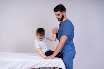The boy sits on a special table, next to him is a doctor in blue uniform and he helps the guy to do physical exercises, rehabilitation concept