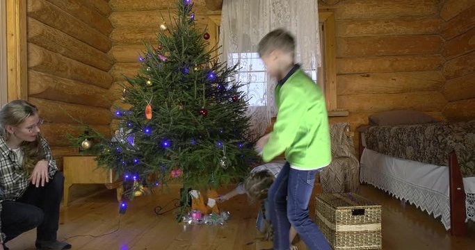 Mother with two children dresses up Christmas tree for the holiday. In the interior of the village house, a mother with children amicably take out Christmas toys and hang them on a green fluffy tree.