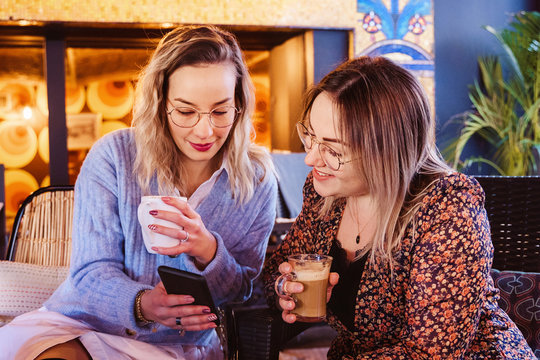Horizontal view of young girlfriends using technology at a restaurant. Couple of women drinking coffee at a terrace on spring holidays and calling a friend with smartphone. - Powered by Adobe