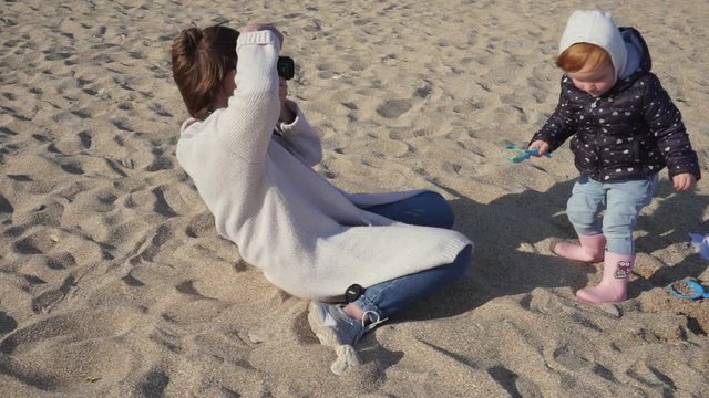 Young and pretty mother photographer is making pictures of her cute red hair daughter on the beach in sand 