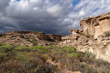 Eroded Volcanic ash and breccia Cliffs rising above the Valley floor covered in Bushes and Succulents, under dark clouds rolling in.