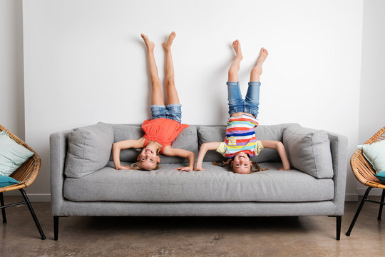 Two smiling young girls doing headstands on sofa at home