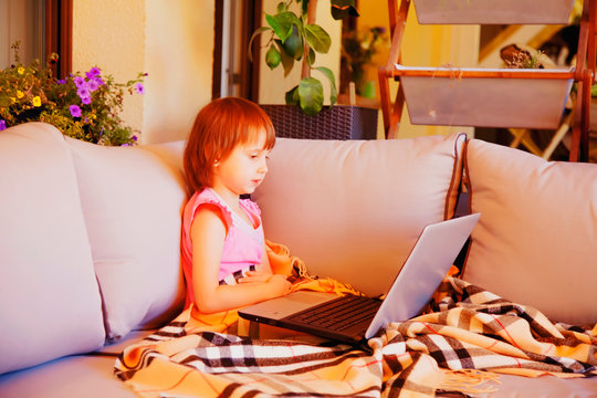 Close up sick cute little child girl siiting on the couch on the porch and studies with laptop remotely. Horizontal image.