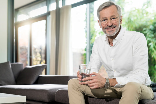 Portrait of smiling senior man in modern design living room sitting on couch holding glass of water