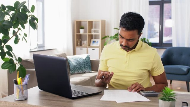 remote job, technology and people concept - young indian man with calculator, laptop computer and papers working at home office