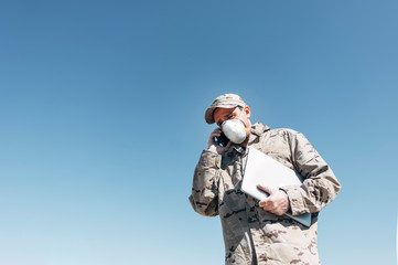 Soldier with face mask on emergency operation, using smartphone, carrying laptop