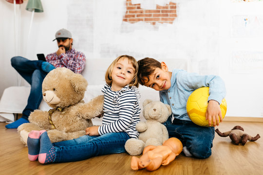 Brother And  Sister Sitting In Playroom With Cuddly Toys, Father In Background