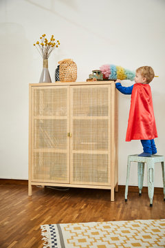 Boy wearing superman costume and cleaning toy with feather duster on a sideboard at home