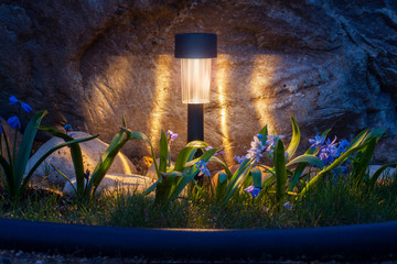 Solar lamp illuminates with blue flowers near a stone in the dark. Garden flashlight shines at...