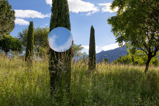Woman hiding behing tree, holding mirror, reflecting the sky