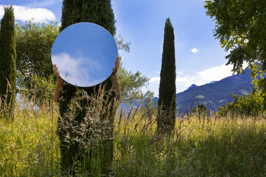 Woman hiding behing tree, holding mirror, reflecting the sky