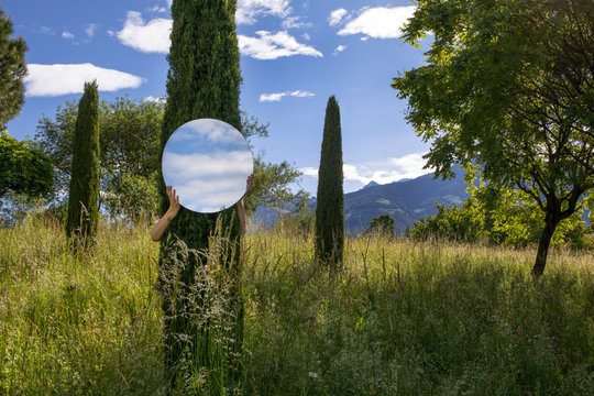 Woman hiding behing tree, holding mirror, reflecting the sky