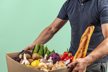 Food delivery, the courier holds a cardboard box with baguette, red pepper, cucumber, lemon, potato, cabbage, garlic, onions. Donation, delivery service.