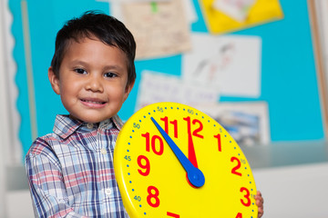Young boy with a preppy shirt, holding a big analog clock with a colorful bulleting board in the background.