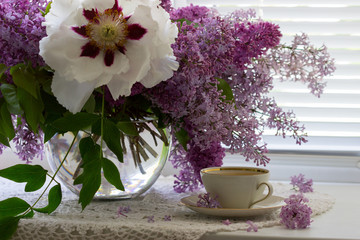 A cup of chamomile tea in a porcelain cup and a bouquet of lilac with peony on a windowsill.