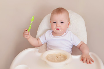 A cheerful child of 10-12 months eats oatmeal with a spoon. Portrait of a happy kid in a highchair.