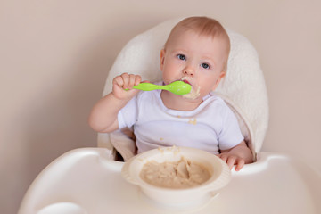 A cheerful child of 10-12 months eats oatmeal with a spoon. Portrait of a happy kid in a highchair.