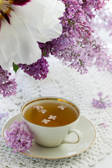 A cup of chamomile tea in a porcelain cup and a bouquet of lilac with peony on a windowsill.