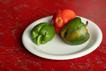 rotten red and green bell peppers on a white plate and a wooden red textured background. Food waste, compost. Old moldy vegetables, horizontal