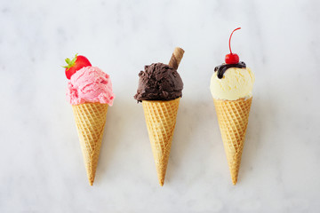 Ice cream cone assortment. Strawberry, chocolate and vanilla in waffle cones. Top view over a white marble background.