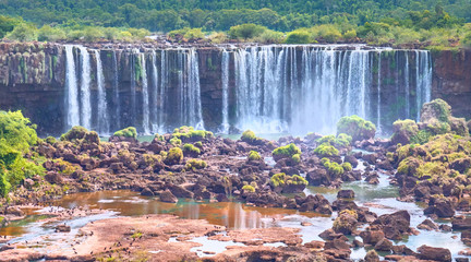 Iguazu waterfalls in Argentina, view from Devil's Mouth, close-up on powerful water streams creating mist over Iguazu river. Sub-tropical foliage in Iguasu river valley, red stone river bed..