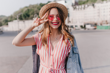 Fashionable white girl in trendy denim jacket posing with peace sign. Cheerful young woman in striped dress and pink sunglasses having fun in city.