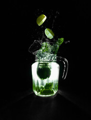 Jarra de mojito iluminada sobre fondo negro, con salpicadura de unas limas cayendo