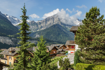 Fototapeta na wymiar Amazing view of the snowy mountain peaks of the Swiss Alps from the small resort village of Champery in Switzerland. Bright blue sky and white fluffy clouds over rocky peaks. Chalets along the road