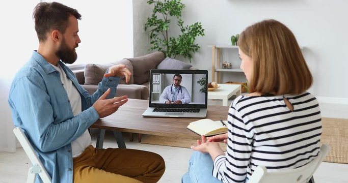 Young man and woman sitting at table and sharing problems with therapist while making video call to psychotherapy clinic