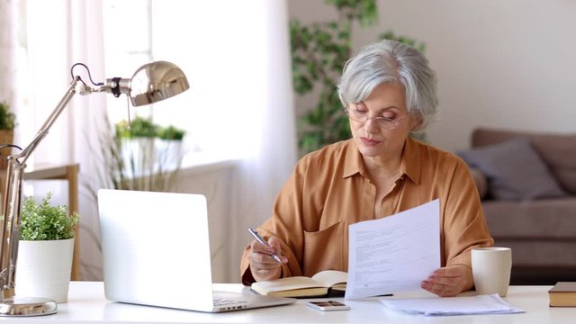 Elderly woman in glasses reading document and making notes in planner while doing remote job in cozy room at home
