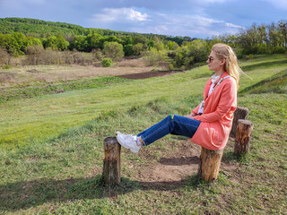 Naklejka na ściany i meble Young woman in colorful clothes sitting on a stump on a background of green hills.