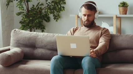  Young man in casual clothes with headphones choosing playlist on laptop and then enjoying music while resting on sofa in cozy living room at home