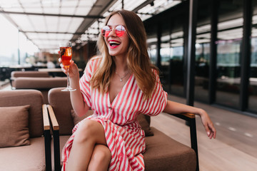 Blissful fair-haired girl spending summer day in restaurant. Happy european woman wears striped dress drinking champagne and smiling.
