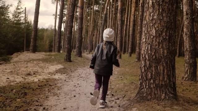 The little girl runs along the path road in the pine forest. Dressed in the leather jacket.