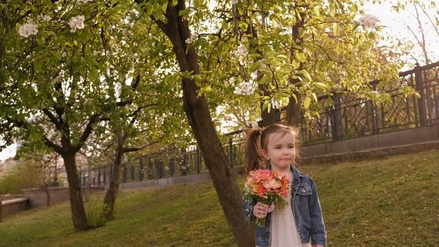 Portrait of a cute charming three-year-old girl with a bouquet of flowers in a spring Park in the sunset sun, she sniffs a bouquet of flowers.