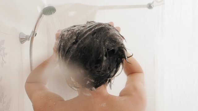 Naked woman takes a shower. Woman washes her short hair with shampoo. Slow motion video in white bathroom.