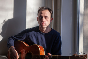 Obraz premium Guy with a guitar in the kitchen. Natural light comes to the musician from the window. Lesson playing the guitar. Unknown talented musician. Playing an acoustic guitar. Brutal man shows skill.