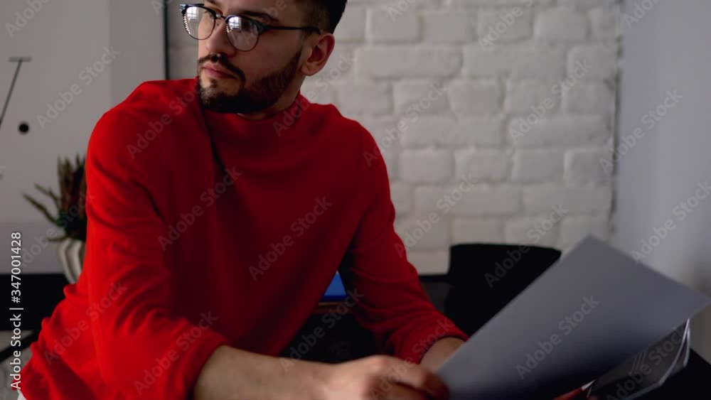 Poster young man dressed in casual wear viewing paper sketches sitting at table and working remotely at hom