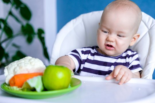 baby food. Little displeased caucasian child pushes plate away from himself with healthy food on highchair. Fresh vegetables and fruits. New born infant kids.