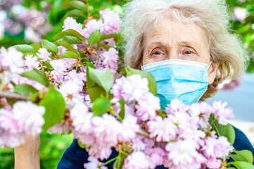 Ederly woman in a protective respiratory mask in a blossoming pink sakura tree. Quarantine, health,...