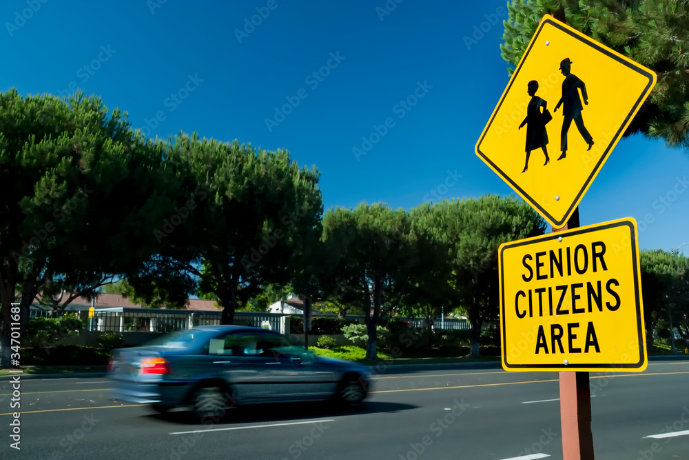 Wall mural Yellow traffic sign that says Senior Citizens Area with graphic of senior citizens walking . Car is blurred driving by in background