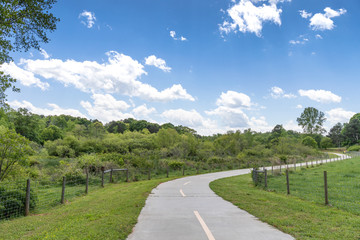 Paved path between meadow and forest, public greenway walking and biking path, blue sky, horizontal aspect