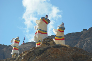 Three shrines made of white fabric rest on top of a rocky outcrop of barren hills in Ladakh. They...